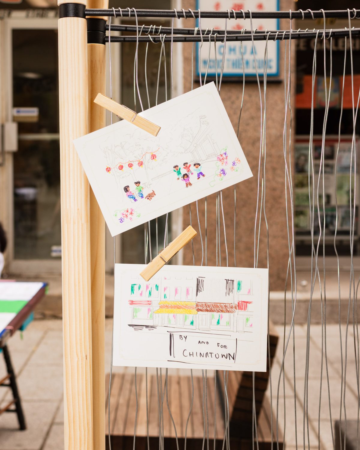 Two postcards pinned onto a makeshift rack. One of them shows smiling children. The other shows colourful buildings, with the words By and For Chinatown.